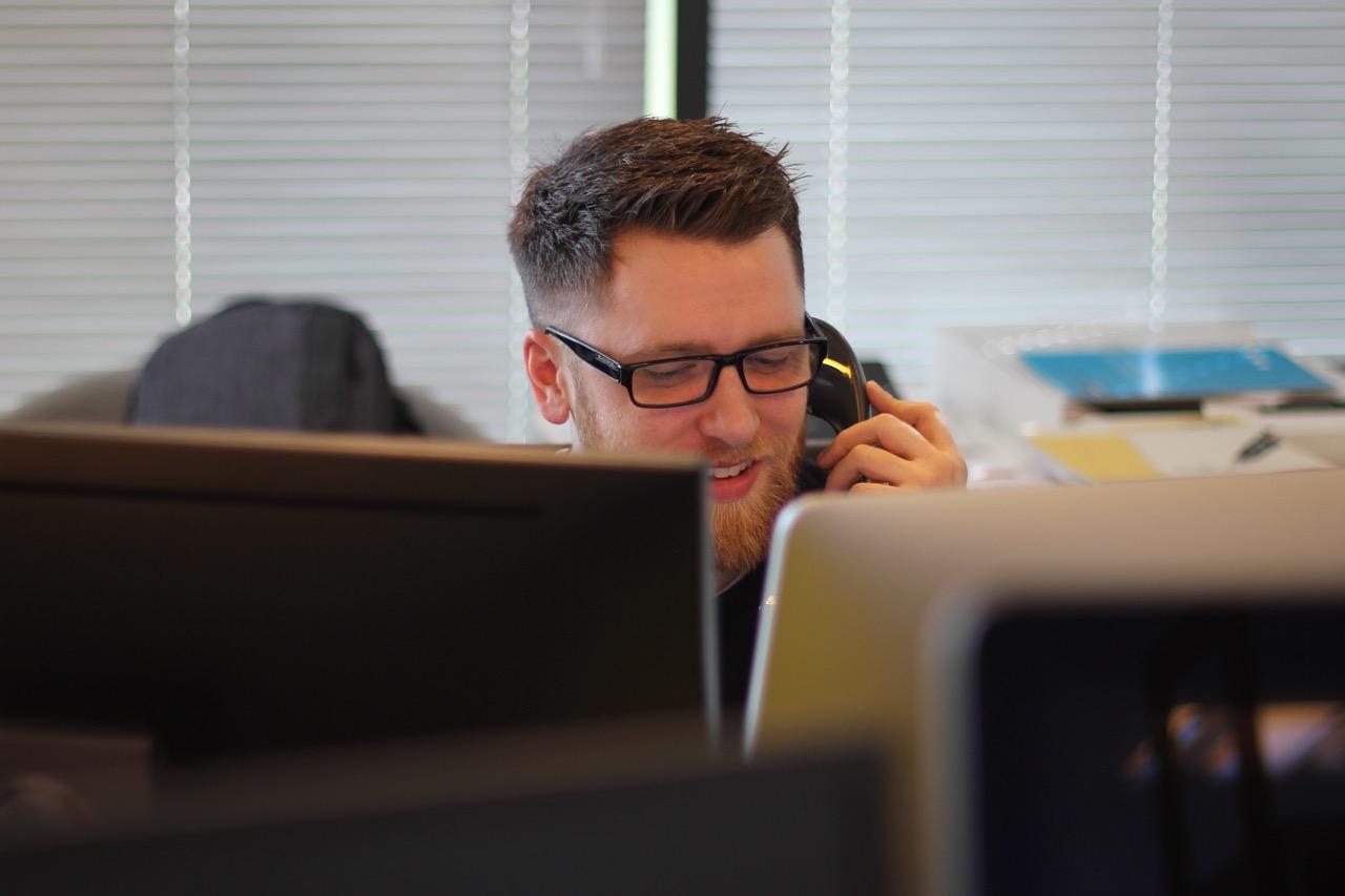A man wearing glasses talking on the phone while sitting in front of a computer.